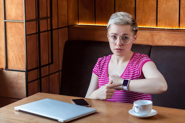 Ritratto di giovane e seria bussineswoman con i capelli corti biondi in maglietta rosa è seduta in un caffè puntando il dito verso l'orologio intelligente da polso e guardando la fotocamera con la faccia triste Stile di vita interno