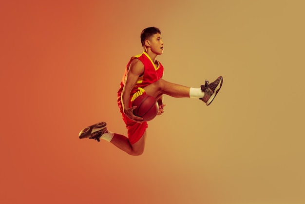 Ritratto di giovane giocatore di basket in movimento che salta formazione isolato sopra lo studio arancione