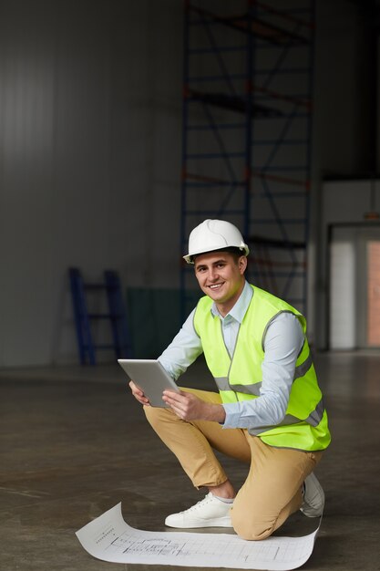 Ritratto di giovane ingegnere nel casco da lavoro sorridendo alla telecamera mentre si lavora con blueprint e tavoletta digitale in cantiere