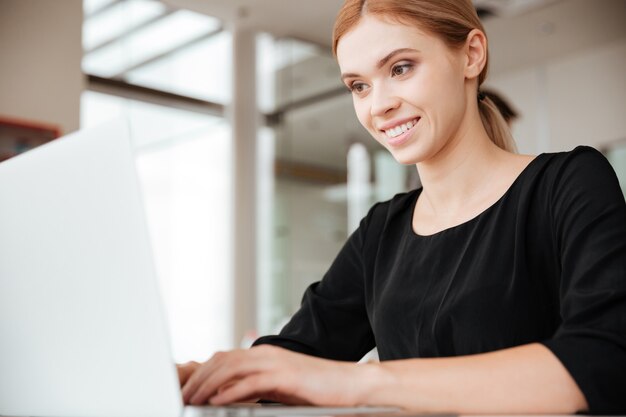 Ritratto di giovane lavoratrice felice seduta in ufficio mentre si utilizza il computer portatile. Guardando il computer.