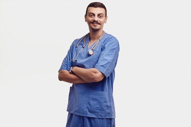 Ritratto di giovane medico arabo sicuro bello con i baffi operati in blu isolato su bianco