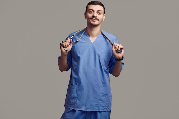 Ritratto di giovane medico arabo sicuro bello con i baffi operati in blu sullo studio grigio