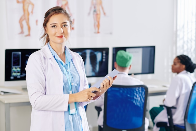 Ritratto di giovane medico generico femminile positivo che tiene computer tablet e sorridente davanti quando si trova in studio medico