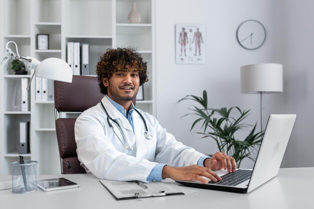 Ritratto di giovane medico ispanico che lavora nell'ufficio con un computer portatile che si siede alla tavola in a