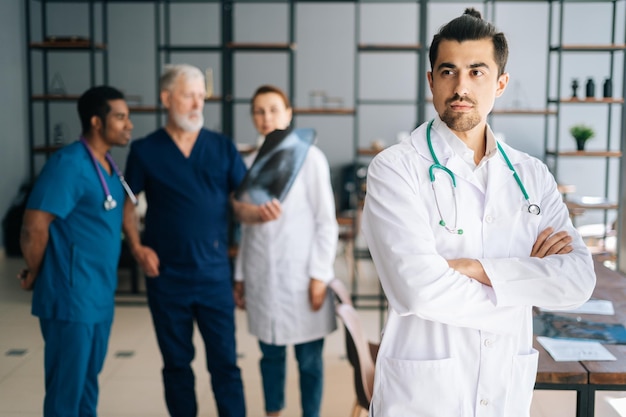 Ritratto di giovane medico maschio fiducioso in uniforme bianca in piedi con le mani incrociate nell'ufficio della riunione medica che distoglie lo sguardo