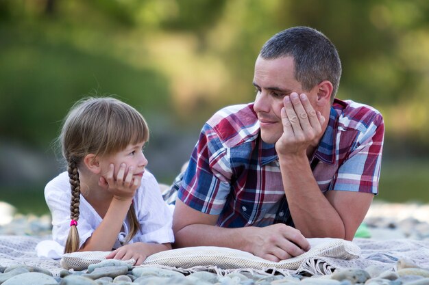 Ritratto di giovane padre e ragazza bionda sveglia con le trecce lunghe che pongono sulla sponda del fiume pebbled il giorno di estate luminoso sugli alberi verdi vaghi. Amore, cura e perfetto concetto di vacanza.