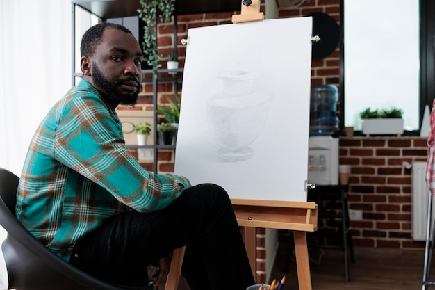 Ritratto di giovane pittore seduto davanti a una tela bianca che disegna un vaso di schizzo usando la matita grafica durante la lezione d'arte nello studio della creatività. Artista sorridente che sviluppa abilità artistiche per la crescita personale