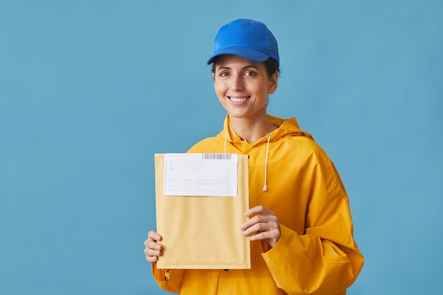 Ritratto di giovane postina in uniforme che tiene la lettera e sorridente sull'azzurro