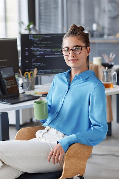 Ritratto di giovane programmatore in occhiali seduto in ufficio e guardando la fotocamera mentre beve il caffè