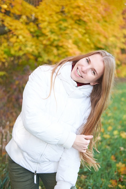 Ritratto di giovane ragazza allegra attraente con bei capelli lunghi naturali indossa giacca bianca e pantaloni verdi. Alberi gialli di autunno su fondo. Tempo d'autunno.