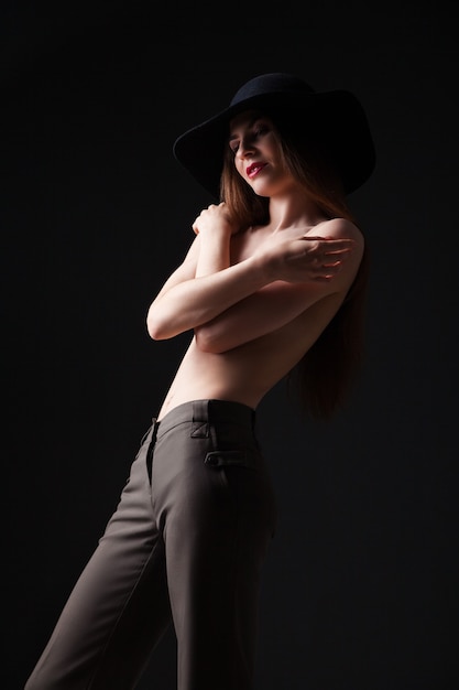 Ritratto di giovane ragazza attraente in cappello nero che copre il seno nudo mentre distoglie lo sguardo. Studio shot