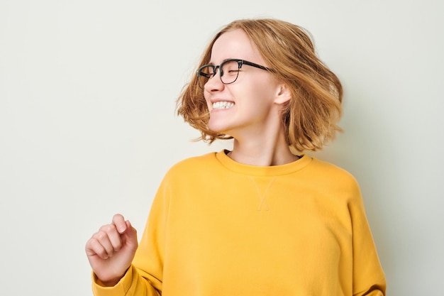 Ritratto di giovane ragazza bionda carina con gli occhiali e maglione giallo scuote la testa e gioca con i capelli isolati su sfondo bianco studio