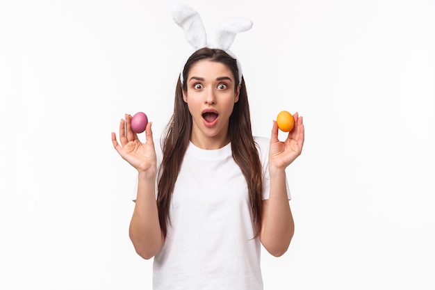 Ritratto di giovane ragazza bruna sorpresa e stupita che tiene due uova colorate, con indosso orecchie di coniglio, vestito da festa per celebrare il giorno di Pasqua