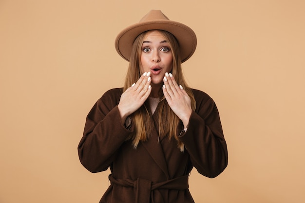 Ritratto di giovane ragazza caucasica che indossa cappello e cappotto esprimendo meraviglia e alzando le mani al viso isolato su beige