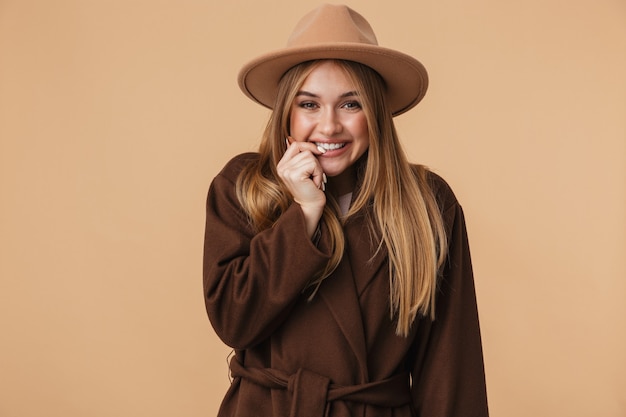 Ritratto di giovane ragazza caucasica che indossa cappello e cappotto sorridente isolato su beige