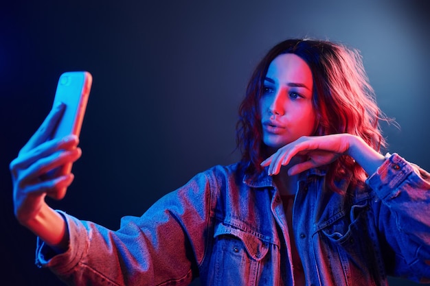 Ritratto di giovane ragazza con i capelli ricci che fa selfie utilizzando il telefono in neon rosso e blu in studio