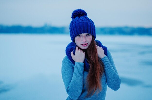 Ritratto di giovane ragazza dai capelli rossi con le lentiggini che indossano un cappello di lana lavorato a maglia blu e una sciarpa nel ghiaccio di sfondo del giorno d'inverno