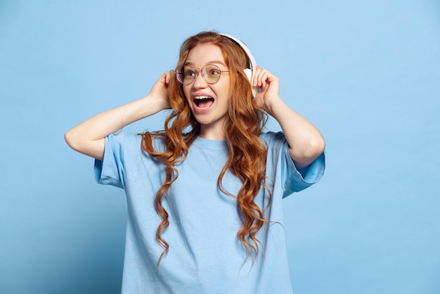 Ritratto di giovane ragazza dai capelli rossi emotiva in cuffia che urla isolato su sfondo blu per studio