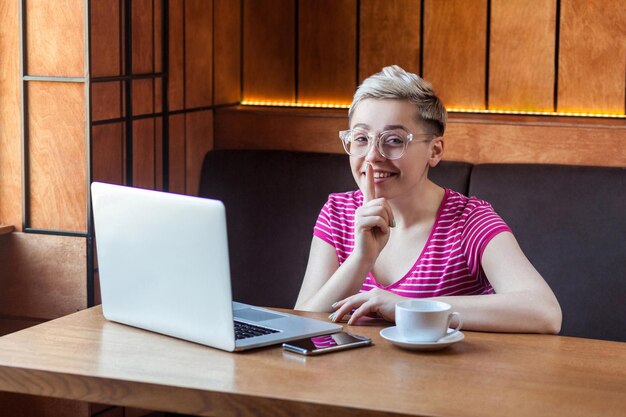 Ritratto di giovane ragazza freelance con i capelli corti in maglietta rosa e occhiali da vista è seduto al bar e si appoggia il dito sulla bocca che mostra il silenzio guardando la fotocamera con un sorriso a trentadue denti