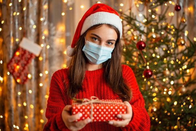 Ritratto di giovane ragazza in maschera medica e cappello santa in piedi davanti all'albero di Natale con un regalo