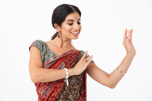 Ritratto di giovane ragazza indù che indossa il tradizionale abito sari indiano e gioielli etnici che ballano con le mani gesticolanti isolate su un muro bianco