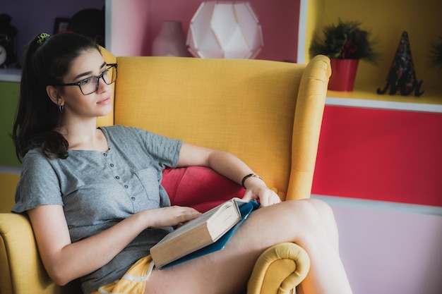 Ritratto di giovane ragazza intelligente e nerd che indossa occhiali che si rilassa sul divano a casa leggendo un libro mentre sorride e guarda la telecamera