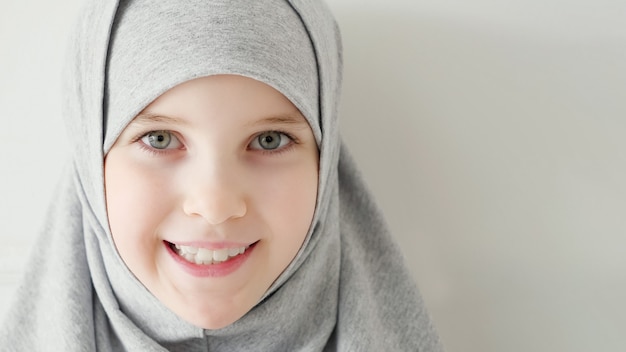 Ritratto di giovane ragazza musulmana attraente di 9 anni che indossa hijab grigio e abito tradizionale guardando la telecamera e sorridente su sfondo chiaro, copia spazio.