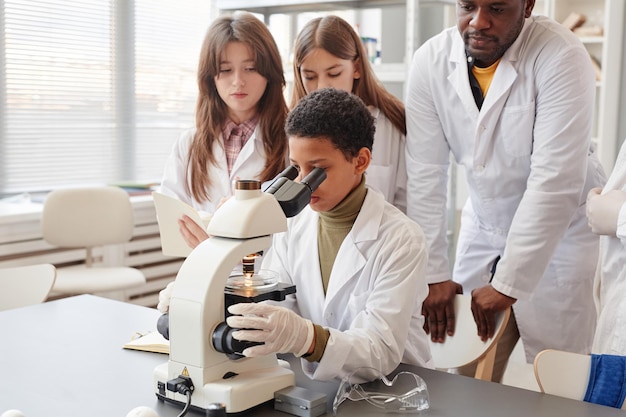 Ritratto di giovane ragazza nera che esamina il microscopio mentre fa esperimenti con un gruppo di bambini nel laboratorio di chimica della scuola