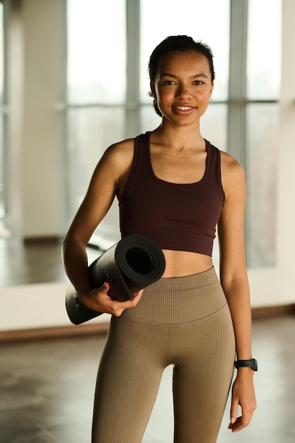 Ritratto di giovane ragazza sana con tappetino per esercizi che sorride alla macchina fotografica mentre si trova in palestra