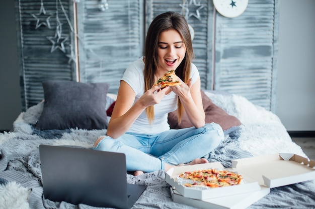 Ritratto di giovane ragazza seduta su un letto con una scatola di pizza, che morde un pezzo di pizza. Donna felice che mangia pizza per colazione.