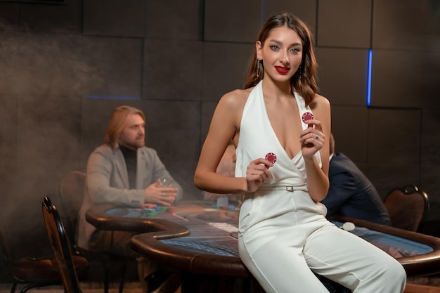 Ritratto di giovane ragazza sorridente che indossa un'elegante tuta bianca con scollo all'americana, tenendo in mano gettoni da gioco rossi, in posa nel casinò appoggiandosi al tavolo da poker con i giocatori che si divertono a giocare in background