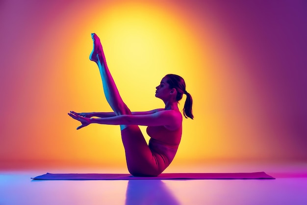 Ritratto di giovane ragazza sportiva che si allena seduta alzando la gamba verso l'alto con le braccia dritte isolate su sfondo rosa e giallo sfumato al neon