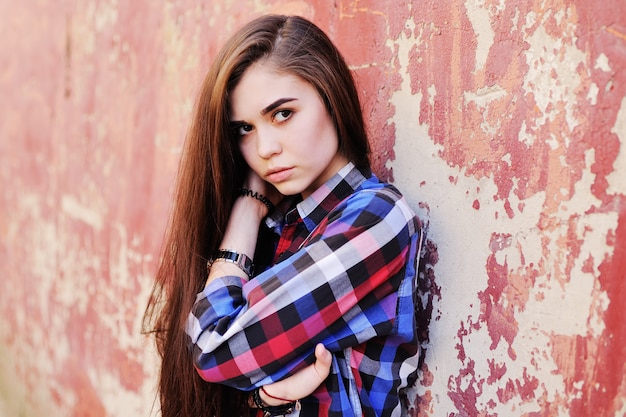 Ritratto di giovane ragazza sveglia del brunette in camicia di plaid sulla superficie rossa della parete del grunge. Copia spazio, spazio per il testo