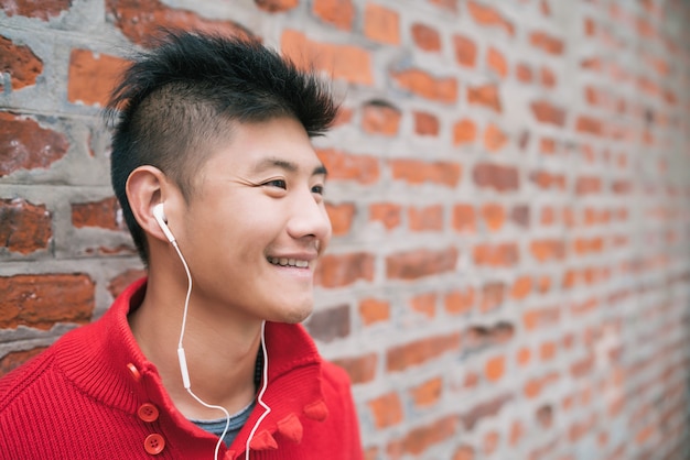 Ritratto di giovane ragazzo asiatico che ascolta la musica con gli auricolari all'aperto contro il muro di mattoni. Concetto urbano.