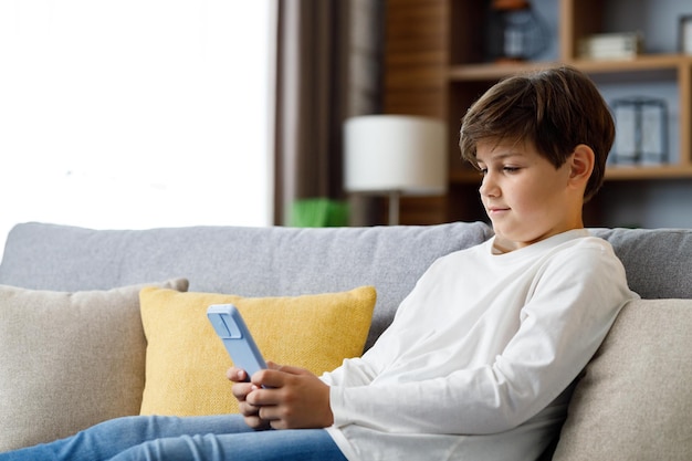 Ritratto di giovane ragazzo carino che tiene smartphone seduto al divano Adolescente a casa che sceglie la musica preferita messaggi di testo navigando in Internet guardando video nei social media giocando a giochi online