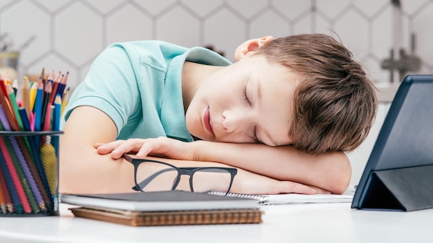 Ritratto di giovane ragazzo stanco del preteen che si trova dormendo sulle mani sulla scrivania vicino ai taccuini occhiali matite colorate tablet
