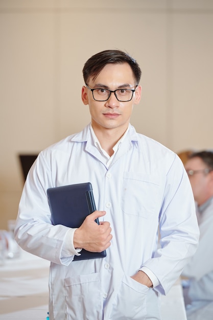 Ritratto di giovane ricercatore fiducioso con gli occhiali in piedi nella sala riunioni con la tavoletta digitale in mano