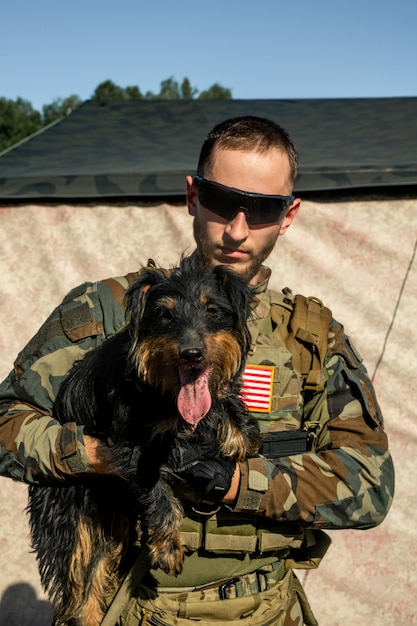 Ritratto di giovane soldato barbuto serio in occhiali da sole e vestito mimetico che tiene un cane carino in un accampamento militare