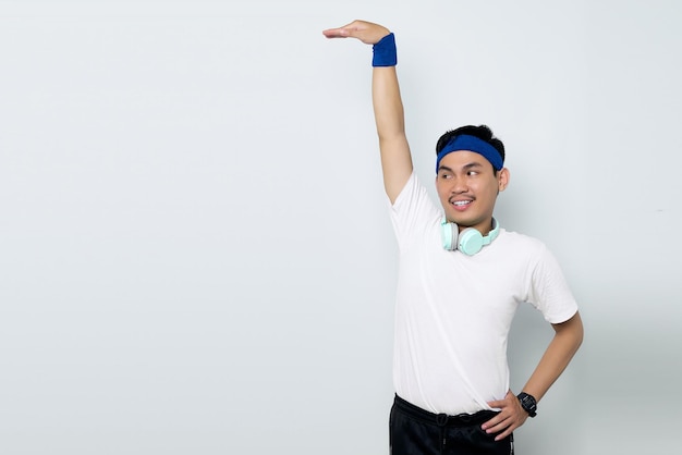 Ritratto di giovane sportivo asiatico sorridente in fascia blu e maglietta bianca da abbigliamento sportivo con le cuffie che mostrano la misura dell'altezza isolata su sfondo bianco Concetto di sport di allenamento
