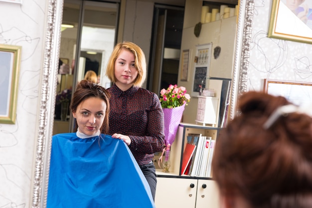 Ritratto di giovane stilista biondo in piedi dietro il giovane cliente femmina bruna che indossa grembiule blu e seduto in poltrona in salone - riflesso nel grande specchio