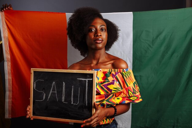 Ritratto di giovane studentessa africana in abiti nazionali che tiene una lavagna con la parola Hello sulla sua lingua madre prima della bandiera della Costa d'Avorio