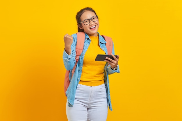 Ritratto di giovane studentessa asiatica allegra in abiti casual con zaino che gioca al videogioco sul telefono cellulare e celebra il successo isolato su sfondo giallo