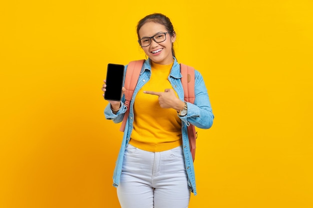 Ritratto di giovane studentessa asiatica allegra in abiti casual con zaino che indica il telefono cellulare con schermo vuoto isolato su sfondo giallo. Istruzione nel concetto di università universitaria