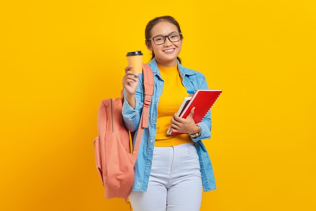 Ritratto di giovane studentessa asiatica allegra in abiti casual con zaino che tiene libro e mostra il caffè da asporto guardando la fotocamera isolata su sfondo giallo