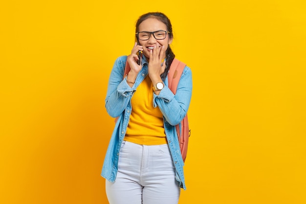 Ritratto di giovane studentessa asiatica allegra in abiti casual con zaino che tiene smartphone e che copre la bocca con le palme isolate su sfondo giallo. Istruzione nel concetto di università universitaria