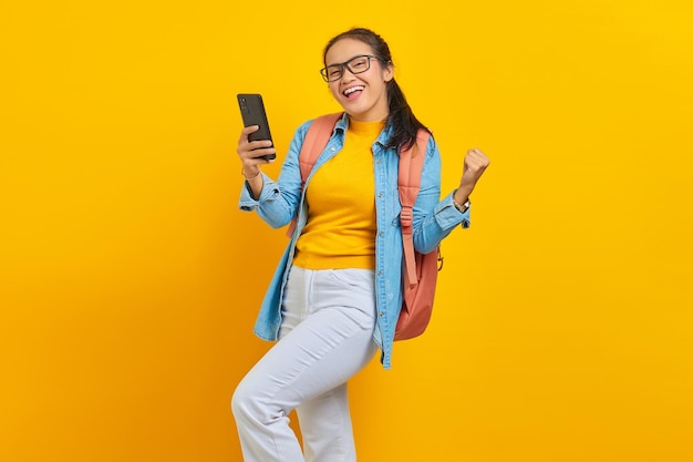 Ritratto di giovane studentessa asiatica allegra in abiti casual con zaino utilizzando il telefono cellulare e celebrando il successo, ottenendo buone notizie isolate su sfondo giallo