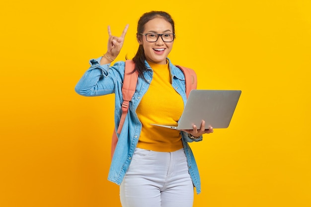 Ritratto di giovane studentessa asiatica allegra in abiti casual con zaino utilizzando laptop e facendo simbolo rock con le mani in alto isolato su sfondo giallo Istruzione nel concetto di college universitario