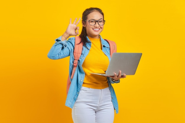 Ritratto di giovane studentessa asiatica allegra in abiti casual con zaino utilizzando laptop e mostrando gesto ok con il dito isolato su sfondo giallo Istruzione nel concetto di college universitario