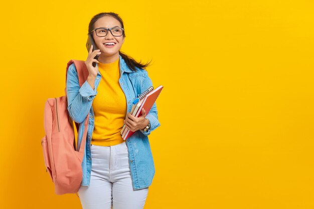 Ritratto di giovane studentessa asiatica allegra in abiti denim con zaino che parla sul telefono cellulare e tiene il libro isolato su sfondo giallo