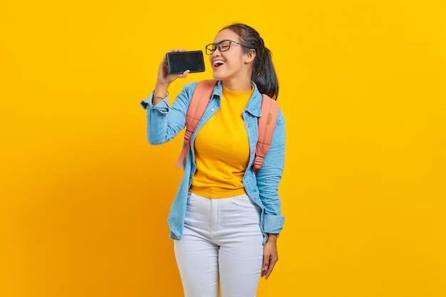 Ritratto di giovane studentessa asiatica sorridente in abiti casual con zaino che si diverte a cantare utilizzando lo smartphone isolato su sfondo giallo. Istruzione nel concetto di università universitaria